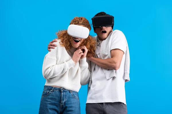 Pareja sorprendida usando auriculares de realidad virtual, aislados en azul - foto de stock