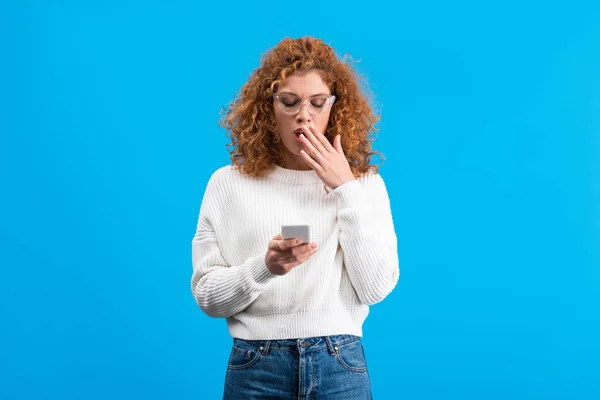 Shocked redhead woman in eyeglasses using smartphone, isolated on blue — Stock Photo