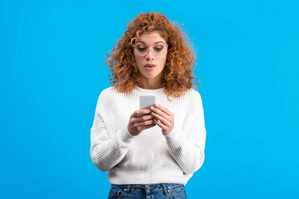 Surprised redhead woman in eyeglasses using smartphone, isolated on blue — Stock Photo