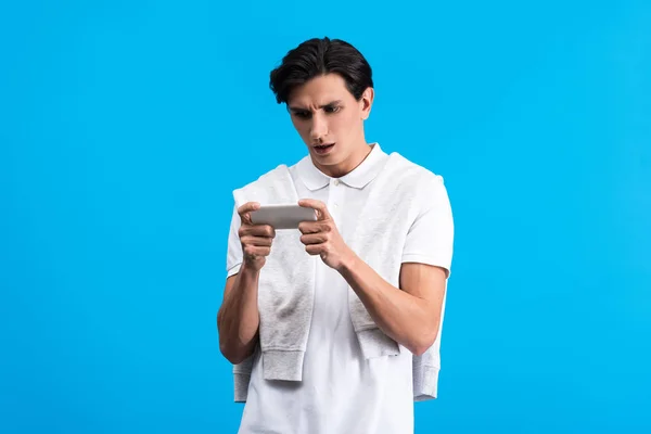 Sorprendido joven usando teléfono inteligente, aislado en azul - foto de stock