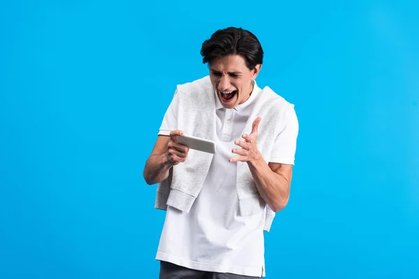 Агрессивный молодой человек кричит и использует смартфон, изолированный на синий — стоковое фото