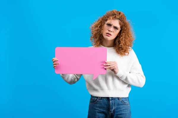 Confused girl in eyeglasses holding empty pink speech bubble, isolated on blue — Stock Photo