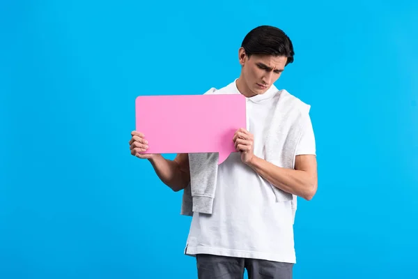 Joven molesto sosteniendo burbuja de habla rosa, aislado en azul - foto de stock