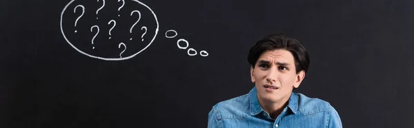 Panoramaaufnahme eines verwirrten jungen Mannes mit Fragezeichen in Gedankenblase an der Tafel — Stockfoto