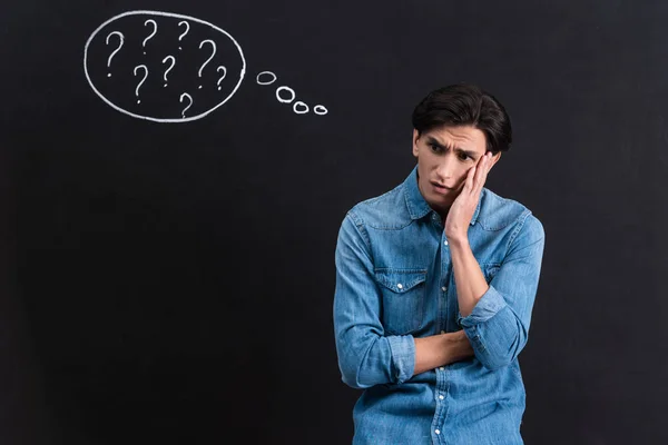 Worried young man with question marks in thought bubble on blackboard — Stock Photo