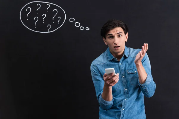 Impactado joven usando teléfono inteligente, con signos de interrogación en el dibujo de la burbuja de pensamiento en pizarra - foto de stock