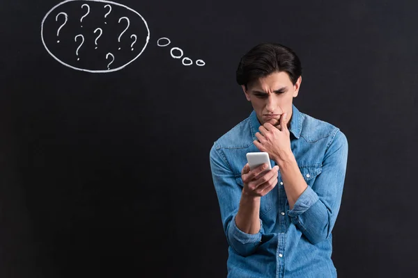 Joven pensativo usando un teléfono inteligente, con signos de interrogación en el dibujo de la burbuja de pensamiento en la pizarra - foto de stock