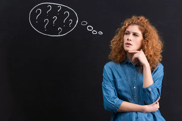 Thoughtful redhead young woman with question marks in thought bubble on blackboard — Stock Photo