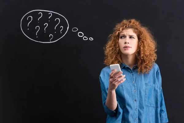 Atractiva mujer pensativa usando smartphone, signos de interrogación en burbuja de pensamiento en pizarra detrás - foto de stock