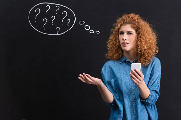 Chica escéptica utilizando el teléfono inteligente, signos de interrogación en burbuja de pensamiento en pizarra detrás - foto de stock