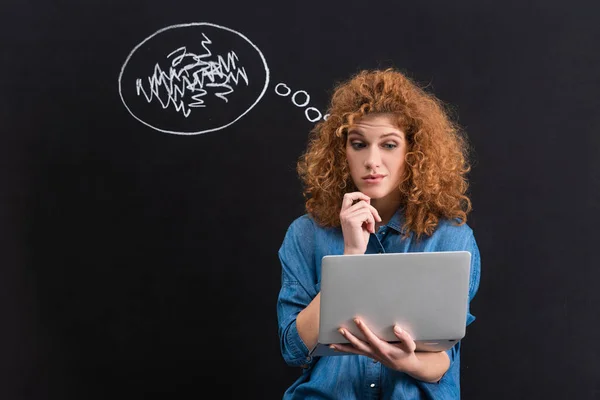 Skeptical redhead girl using laptop with thought bubble on blackboard — Stock Photo