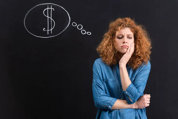 Sad redhead girl with dollar sign in thought bubble on chalkboard — Stock Photo