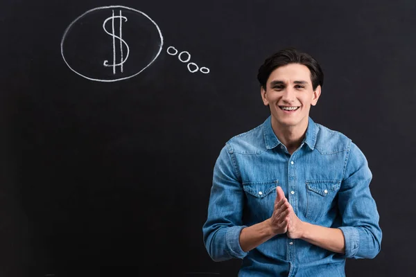 Jeune homme positif avec dollar en signe de bulle de pensée sur tableau noir — Photo de stock