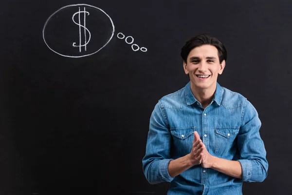 Jeune homme positif avec dollar en signe de bulle de pensée sur tableau noir — Photo de stock