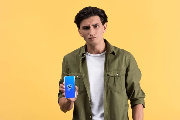 KYIV, UCRANIA - 18 de noviembre de 2019: hombre escéptico mostrando el teléfono inteligente con la aplicación shazam en la pantalla, aislado en amarillo - foto de stock