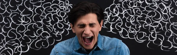Panoramic shot of angry young man yelling with steam drawing on blackboard behind — Stock Photo
