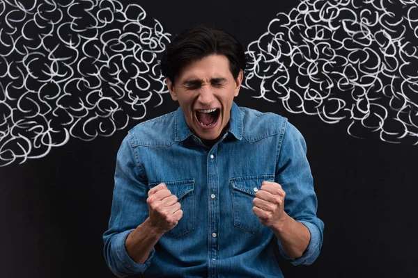 Angry young man holding fists and shouting with steam drawing on blackboard behind — Stock Photo