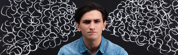 Panoramic shot of irritated young man with steam drawing on blackboard behind — Stock Photo
