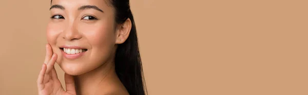 Sonriendo hermosa chica asiática tocando la cara aislado en beige, tiro panorámico - foto de stock