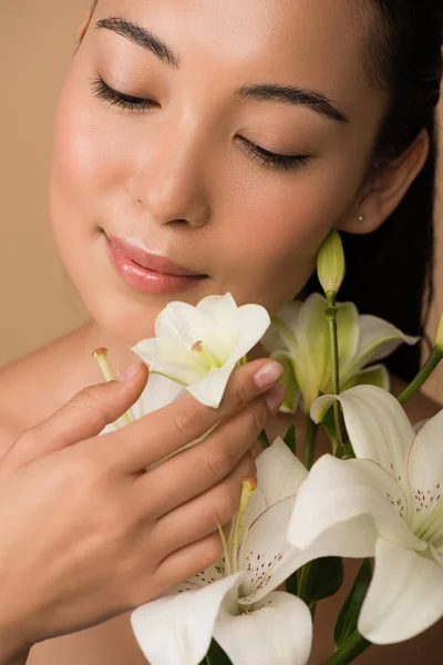 Улыбающаяся красивая обнаженная азиатская девушка с закрытыми глазами и белыми лилиями, изолированными на бежевом — стоковое фото