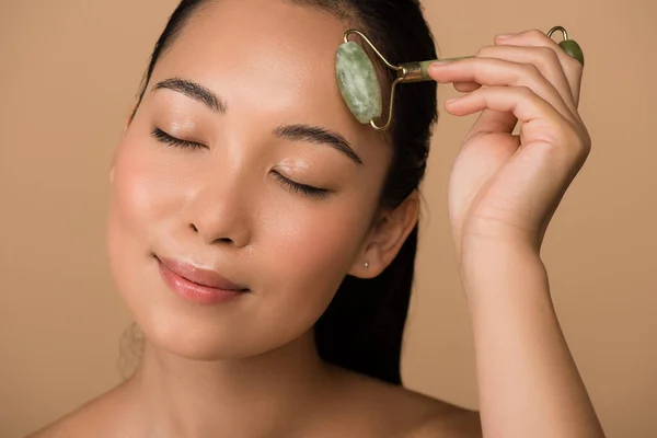 Smiling beautiful naked asian girl with closed eyes massaging face with jade roller isolated on beige — Stock Photo