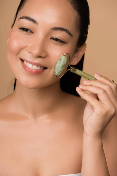 Smiling beautiful naked asian girl massaging face with jade roller isolated on beige — Stock Photo