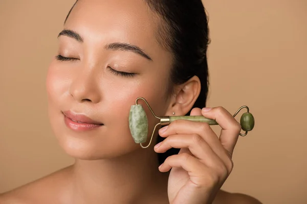 Smiling beautiful naked asian girl with closed eyes massaging face with jade roller isolated on beige — Stock Photo