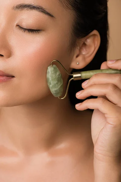 Close up view of beautiful naked asian girl massaging face with jade roller isolated on beige — Stock Photo