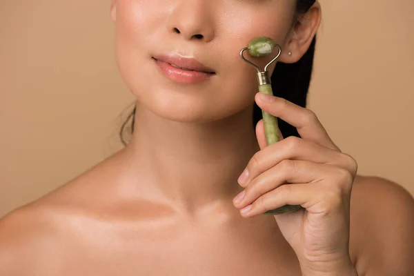 Cropped view of beautiful naked asian girl massaging face with jade roller isolated on beige — Stock Photo