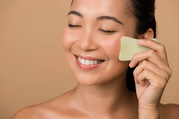 Sonriendo hermosa chica asiática desnuda con los ojos cerrados usando facial gua sha jade board aislado en beige - foto de stock