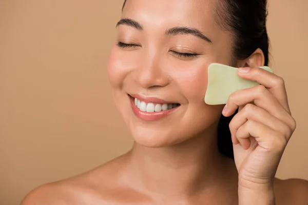 Sorrindo bonito nu asiático menina com olhos fechados usando facial gua sha jade board isolado no bege — Fotografia de Stock