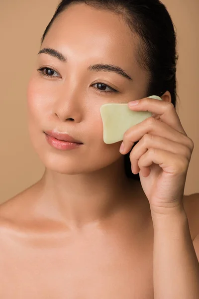 Beautiful naked asian girl using facial gua sha jade board isolated on beige — Stock Photo