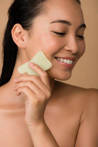 Sonriendo hermosa chica asiática desnuda con los ojos cerrados usando facial gua sha jade board aislado en beige - foto de stock