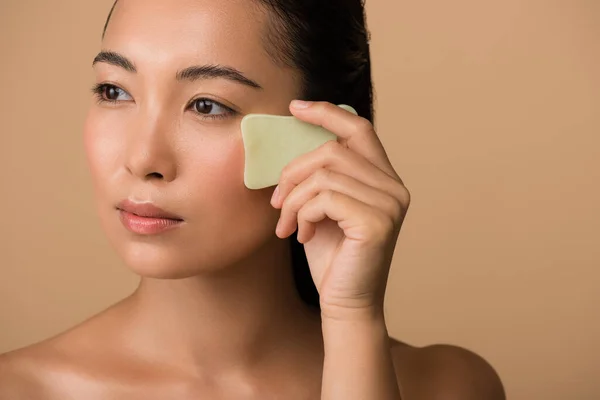 Beautiful naked asian girl using facial gua sha jade board isolated on beige — Stock Photo