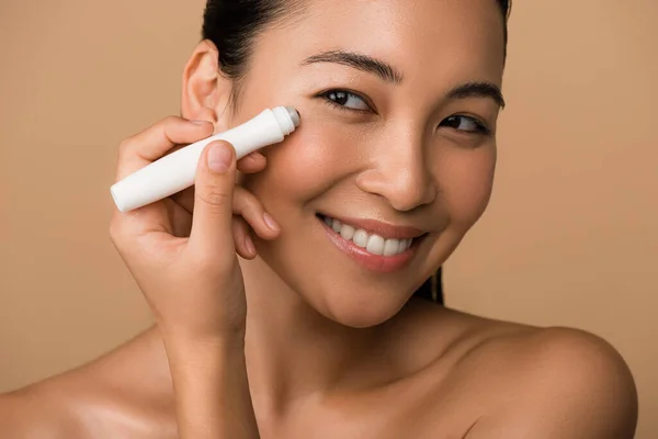 Sonriendo hermosa desnuda chica asiática usando ojo rodillo aislado en beige — Stock Photo