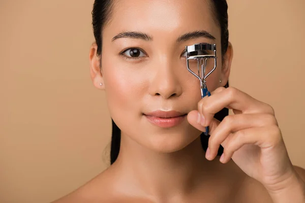 Beautiful naked asian girl using eyelash curlers isolated on beige — Stock Photo