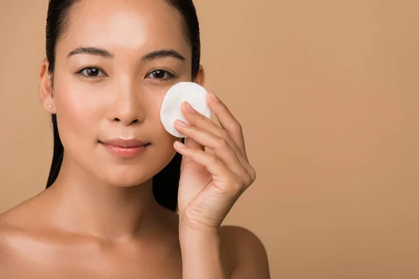 Hermosa chica asiática desnuda sosteniendo almohadilla de algodón en la cara aislado en beige — Stock Photo