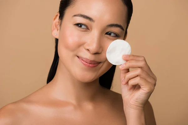 Smiling beautiful naked asian girl holding cotton pad on face isolated on beige — Stock Photo