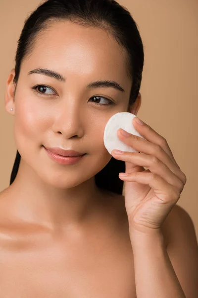 Smiling beautiful naked asian girl holding cotton pad on face isolated on beige — Stock Photo