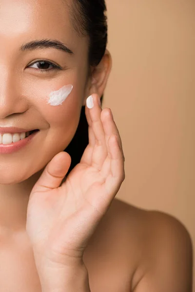 Cropped view of happy beautiful naked asian girl applying facial cream isolated on beige — Stock Photo
