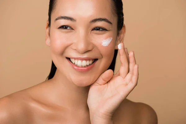 Happy beautiful naked asian girl applying facial cream isolated on beige — Stock Photo