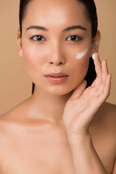Beautiful naked asian girl applying facial cream isolated on beige — Stock Photo