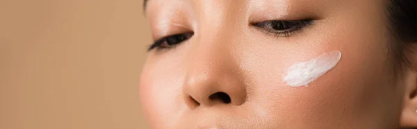 Partial view of beautiful asian girl with facial cream on cheek isolated on beige, panoramic shot — Stock Photo