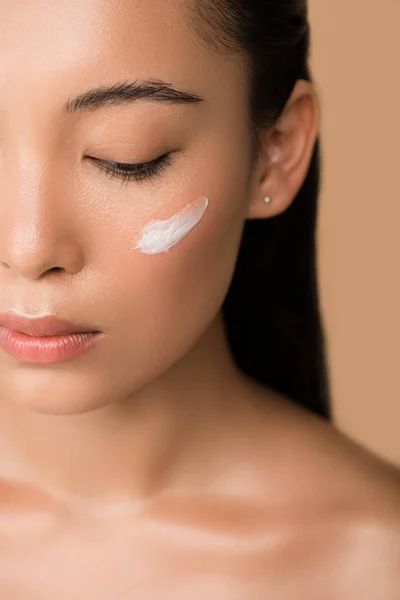 Recortado vista de hermosa chica asiática desnuda con crema facial en la mejilla aislado en beige - foto de stock