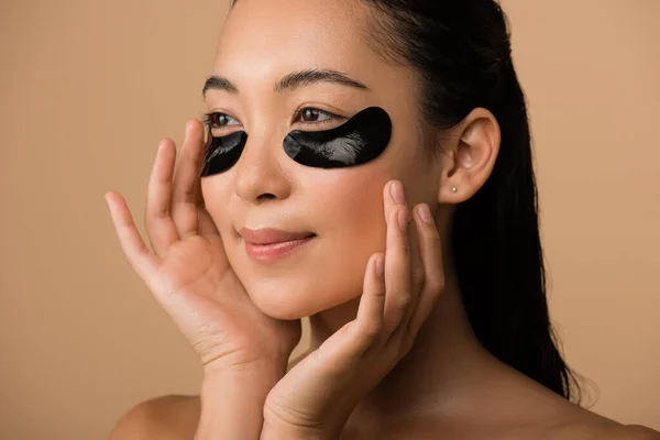 Smiling beautiful naked asian girl with black hydrogel eye patches under eyes isolated on beige — Stock Photo