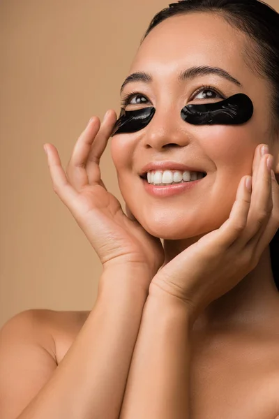 Sonriendo hermosa chica asiática desnuda con parches de hidrogel negro bajo los ojos aislados en beige - foto de stock