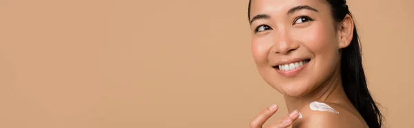 Sorridente bella ragazza asiatica nuda con crema cosmetica sulla spalla isolata su beige, colpo panoramico — Foto stock