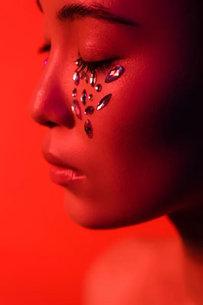 Profile of beautiful asian girl with rhinestones on face and closed eyes isolated on red — Stock Photo