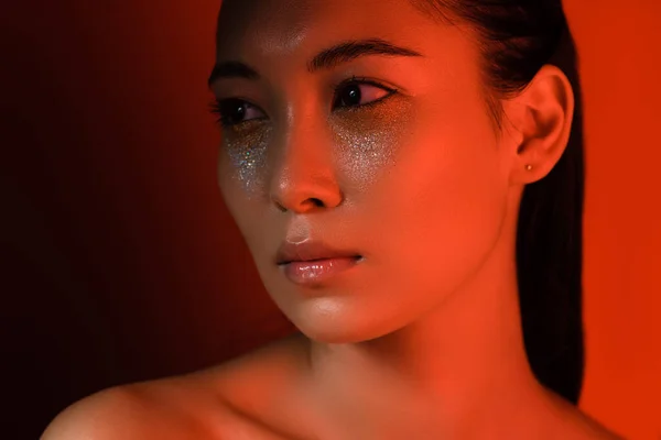 Beautiful naked asian girl with silver sparkles on face in red lighting — Stock Photo