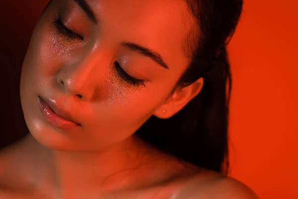 Beautiful naked asian girl with silver sparkles on face and closed eyes in red lighting — Stock Photo